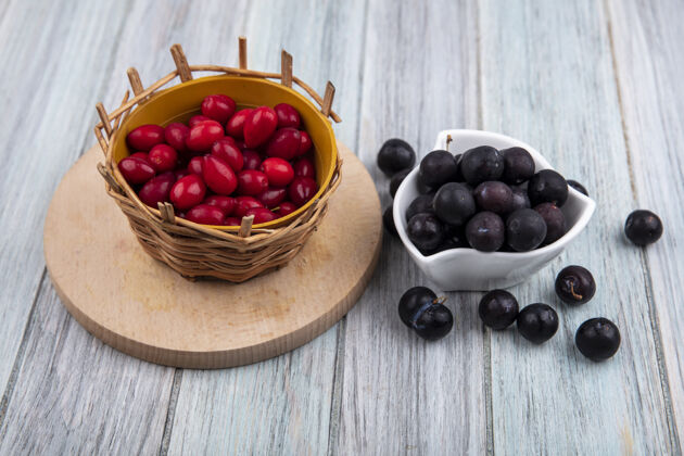 视图白色碗上的小酸黑刺的顶视图 桶上有红色的山茱萸浆果 在灰色木制背景上的木制厨房板上黑刺浆果季节