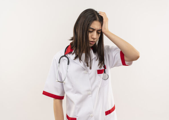 年轻人年轻的女医生穿着白大褂 脖子上戴着听诊器 站在白墙上 看上去很疲惫 而且工作过度看周围人