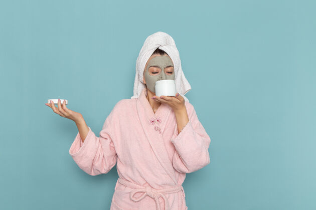 盖正面图身着粉色浴袍的年轻女性拿着面霜在蓝色墙壁上美丽的水浴面霜自理淋浴前面浴袍保护盖