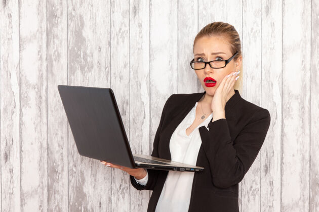 使用正面图穿着严苛衣服的年轻女商人黑色夹克在白墙上使用笔记本电脑工作办公室女商人衣服笔记本电脑女性