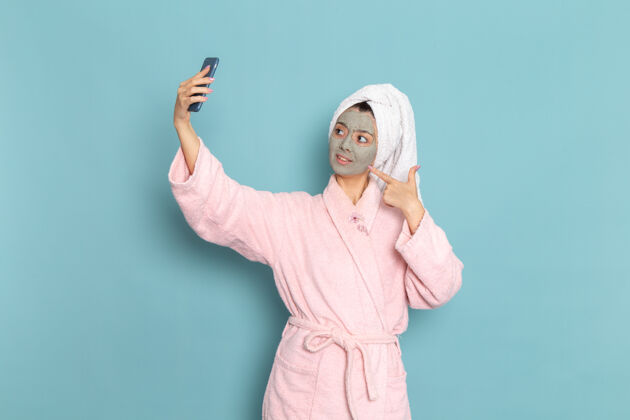 人正面图身着粉色浴袍的年轻女性在蓝色办公桌上自拍 脸上戴着口罩清洁美容自护霜淋浴采取清洁人