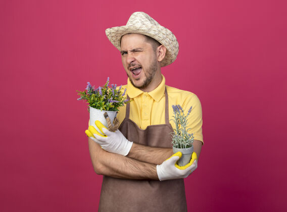 植物年轻的园丁穿着连体衣 戴着帽子 戴着工作手套 手里拿着盆栽植物 站在粉色的墙上微笑着眨眼看着前方花园制服男