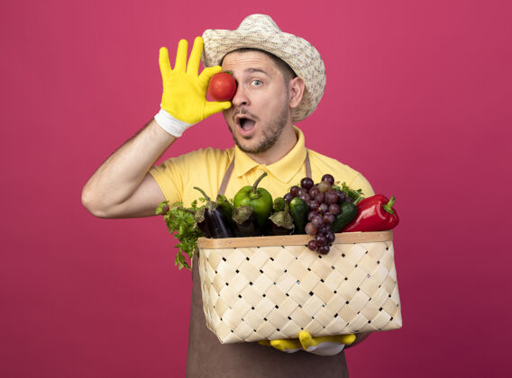 看年轻的园丁穿着连体衣 戴着工作手套 手里拿着装满蔬菜和新鲜西红柿的箱子 站在粉红色的墙上显得很惊讶持有帽子男性