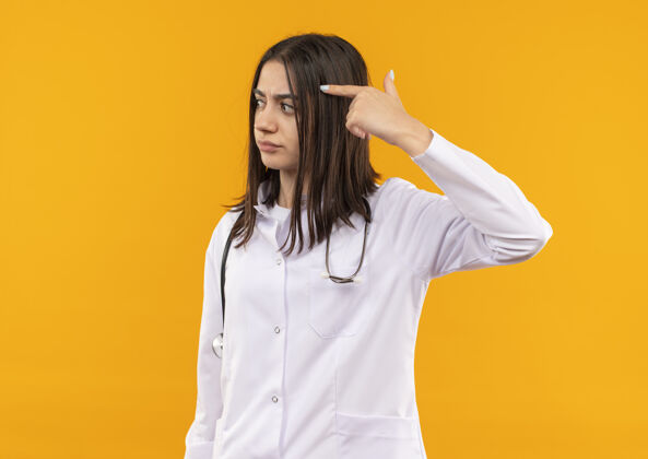 指点身穿白大褂的年轻女医生脖子上戴着听诊器 手指指着太阳穴 站在橘色的墙上专注于一项任务人工作办公室