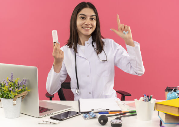 听诊器年轻的女医生穿着白大褂 脖子上戴着听诊器 显示着灯泡和食指有了新的好主意 微笑着坐在桌旁 笔记本电脑盖在粉红色的墙上显示外套医生