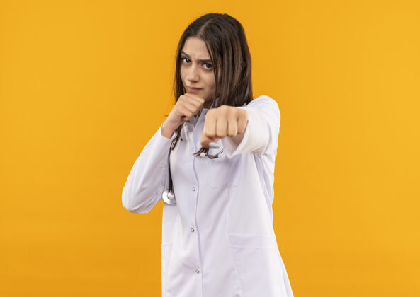 周围年轻的女医生穿着白大褂 脖子上戴着听诊器 表情严肃 站在橙色的墙上像个拳击手站姿势女