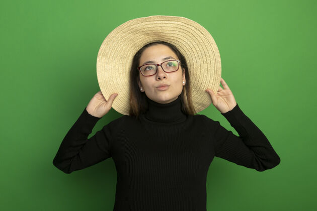 人年轻漂亮的女人戴着夏天的帽子 穿着黑色高领毛衣 戴着眼镜 站在绿色的墙上 高兴而积极地抬头看快乐漂亮女人