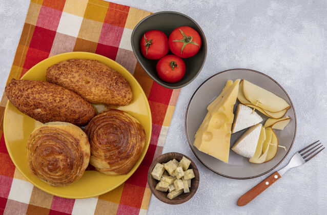 碗格子布上黄色盘子上的新鲜馒头俯视图 灰色盘子上有不同类型的奶酪 碗上有西红柿 背景为白色传统灰色奶酪