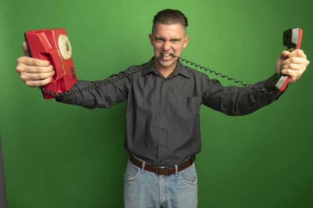 绳子愤怒的年轻帅哥 穿着灰色衬衫 手里拿着老式的电话线 站在绿色的墙上咬表情衬衫