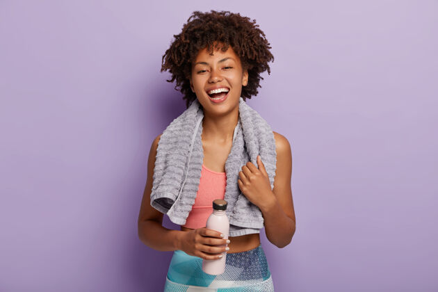出汗身着上衣和紧身裤的微笑健身女士在训练结束后休息 拿着一瓶水 用毛巾擦汗瓶子耐力健康