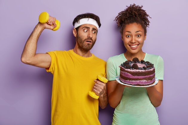 高兴运动健康的生活方式反对垃圾食品困惑的运动员举着哑铃肌肉满意健身房