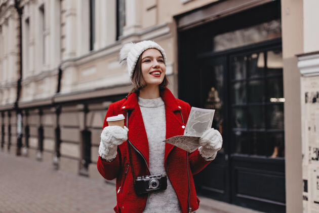 肖像穿着红色保暖夹克 羊绒衫 戴着手套的白帽子的积极的女人拿着咖啡在城市里走来走去脖子上挂着复古相机的游客拿着地图年轻衣服时尚