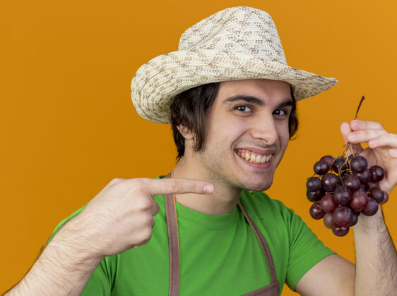 站围裙戴帽子的年轻园丁站在橘红色的墙上 用食指指着一束葡萄 脸上洋溢着幸福的笑容帽子束葡萄