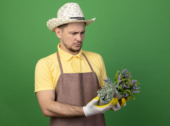 年轻人年轻的园丁穿着连体衣 戴着帽子 戴着工作手套 手里拿着盆栽植物 站在绿色的墙上严肃地看着它们制服锅绿色