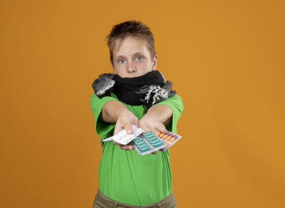 温暖生病的小男孩穿着绿色t恤 脖子和嘴上围着温暖的围巾 站在橘色的墙上 他看起来很担心看小周围
