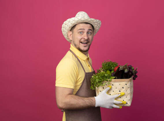 站着年轻的园丁穿着连体衣 戴着帽子 戴着工作手套 手里拿着装满蔬菜的箱子 站在粉色的墙上 面带微笑地看着前方穿脸板条箱