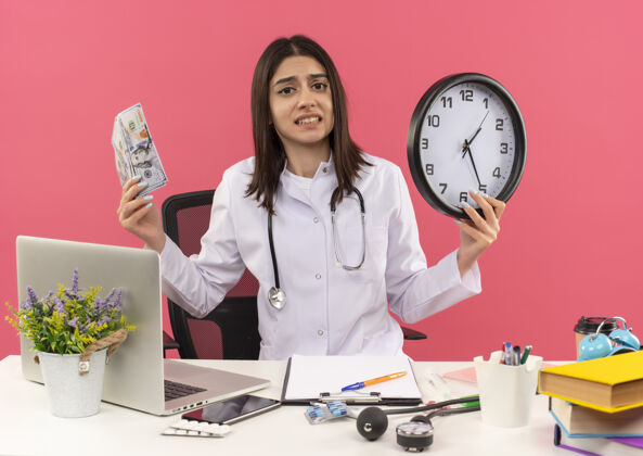 困惑年轻的女医生穿着白大褂 脖子上戴着听诊器 手里拿着现金和挂钟 坐在桌旁 笔记本电脑盖在粉红色的墙上 看上去既困惑又焦虑女人医生脖子