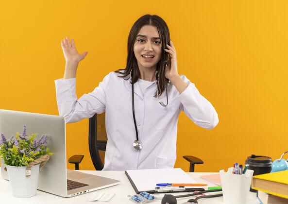 医生年轻的女医生穿着白大褂 手持听诊器讲着手机 坐在桌子旁 手里拿着笔记本电脑 橘黄色的墙上挂着文件 看上去很困惑桌子笔记本电脑年轻