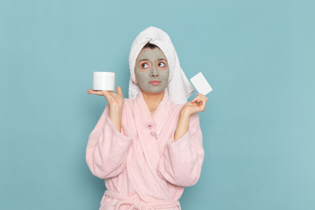 长袍正面图身穿粉色浴袍 脸上戴着面具的年轻女性手持蓝色墙壁上的卡片淋浴清洁美容自护霜面膜保持淋浴