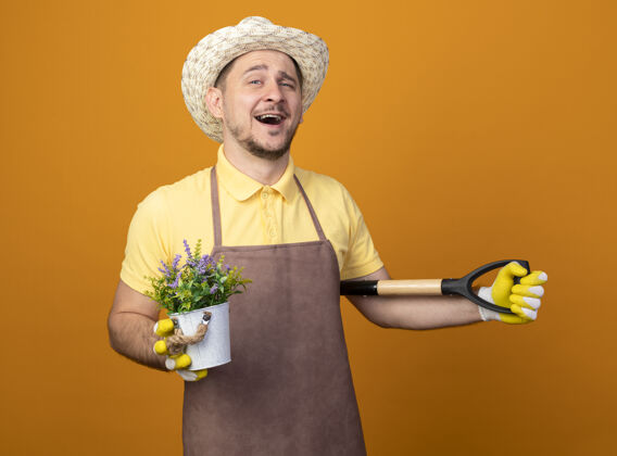 人年轻的园丁穿着连体衣 戴着帽子 拿着铲子和盆栽植物 站在橙色的墙上 面带微笑地看着前方人帽子站着