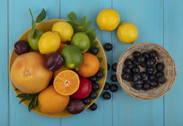盘子顶视图樱桃李子放在一个篮子里 橘子李子柠檬和酸橙放在绿松石背景的黄色盘子上水果樱桃食物