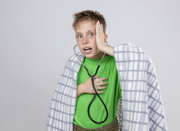 疾病生病的小男孩穿着绿色t恤 裹着毛毯 脖子上戴着听诊器 站在白色的墙上听着心跳混乱包裹听诊器不适