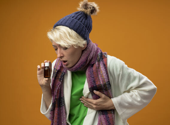暖生病的不健康的女人 短头发 戴着暖和的围巾 戴着不舒服的帽子 拿着感冒药站在橙色的墙上感冒不适橘子