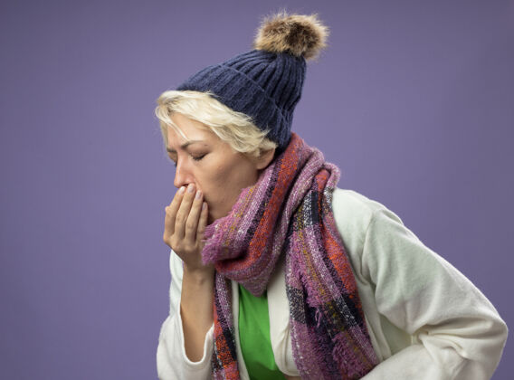 痛苦生病的不健康的女人 短发 戴着暖和的围巾和帽子 站在紫色的墙上 感觉不舒服 咳嗽 流感咳嗽女人感觉