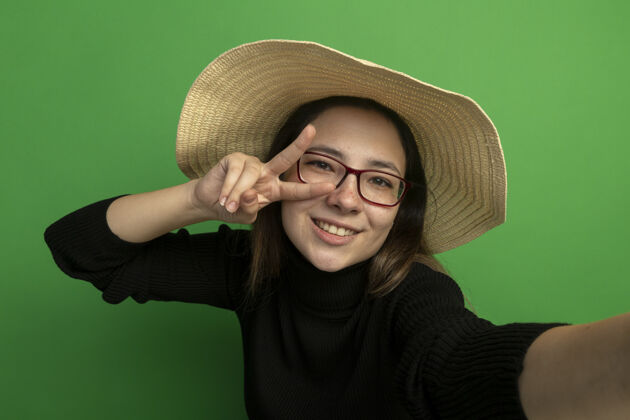 快乐年轻漂亮的女人戴着夏天的帽子 穿着黑色高领毛衣 戴着眼镜 看着前面 快乐而积极的微笑 兴高采烈地做着v形标志站在绿色的墙上高领毛衣使年轻