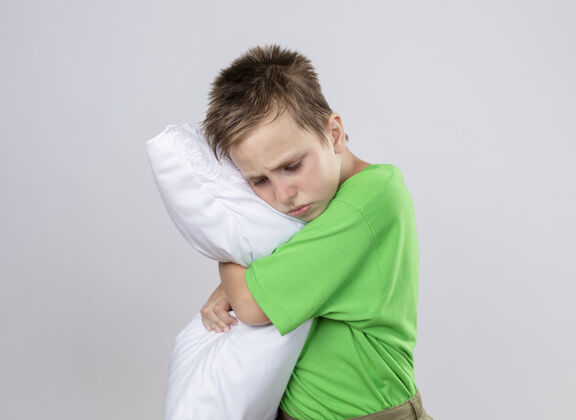 疾病生病的小男孩穿着绿色t恤感觉不舒服抱枕闭着眼睛站在白色的墙上小拥抱枕头