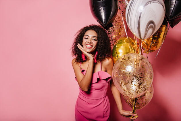 节日迷人的女人微笑着在粉红色的墙上摆姿势气球生日派对时尚