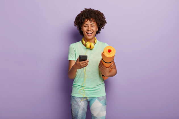 T恤锻炼和健身概念开朗的黑皮肤女人拿着连接耳机的手机护理移动大笑