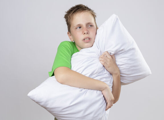站生病的小男孩穿着绿色t恤 感觉不舒服 抱着枕头 站在白色的墙上 看着一边 不舒服 生病病了枕头感觉