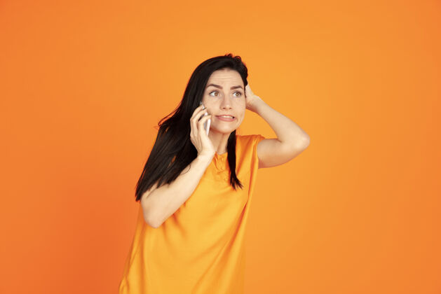 公司橙色工作室背景上的白种人年轻女子肖像穿着衬衫的漂亮黑发女性模特人类情感的概念 面部表情 销售 广告广告空间电话交谈霓虹灯衬衫女性