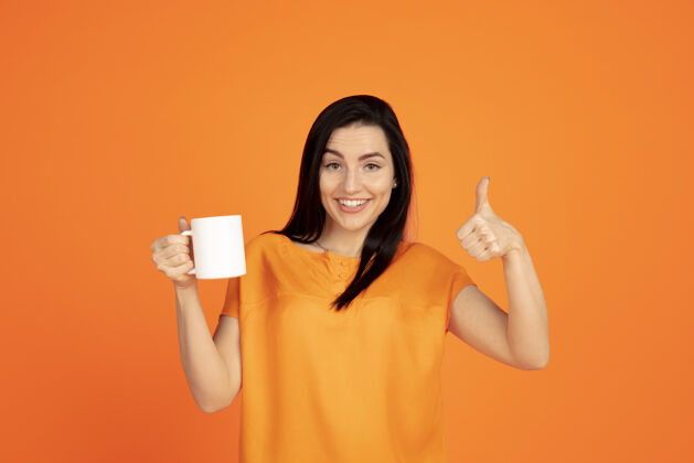 工作橙色工作室背景上的白种人年轻女子肖像穿着衬衫的漂亮黑发女性模特人类情感的概念 面部表情 销售 广告广告空间喝咖啡或茶成功财务面部