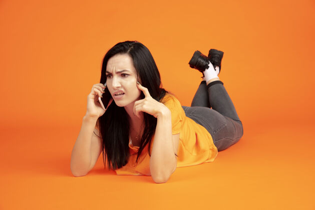 女商人橙色工作室背景上的白种人年轻女子肖像穿着衬衫的漂亮黑发女性模特人类情感的概念 面部表情 销售 广告广告空间电话交谈年轻面部员工