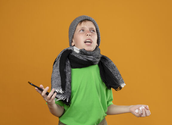餐巾生病的小男孩穿着绿色t恤 戴着暖和的围巾和帽子 感觉不舒服 拿着智能手机和纸巾 站在橙色的墙上 困惑和不高兴地看着一边温暖不舒服围巾