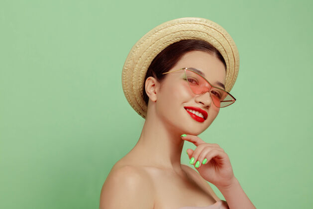 保持美丽女人的肖像 明亮的妆容 帽子和太阳镜在绿色的工作室背景上时髦和时尚的制作和发型夏天的颜色美丽 时尚和广告概念微笑严肃护理自信