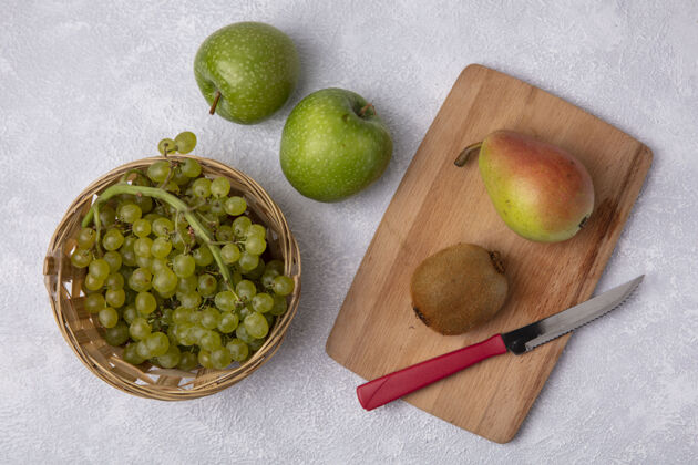 刀顶视图绿色的葡萄在一个篮子里 梨猕猴桃和刀子放在砧板上 绿色的苹果在白色的背景上绿色板猕猴桃