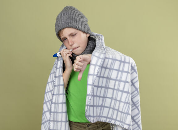 温度生病的小男孩穿着绿色t恤 戴着暖和的围巾 帽子裹着毯子 把温度计放进嘴里 测量温度 拇指朝下站在明亮的墙上男孩毯子穿