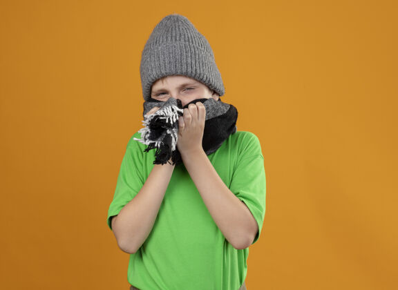 温暖生病的小男孩穿着绿色t恤 戴着暖和的围巾 戴着帽子 戴着围巾 站在橙色的墙上感觉不舒服盖站立少