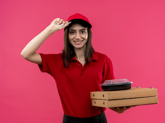 看穿着红色马球衫 戴着帽子的年轻送货员 手里拿着披萨盒和食品包 脸上挂着幸福的笑容 站在粉红色的墙上 摸着帽子站着盒子送货