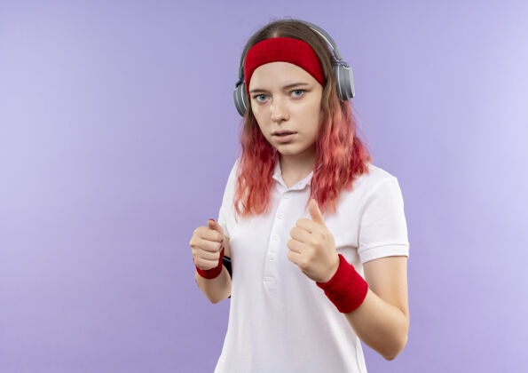 人戴着头戴式耳机 表情严肃的年轻运动女性 手持智能手机袖标站在紫色墙壁上训练训练女运动装