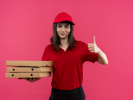 披萨身穿红色马球衫 头戴鸭舌帽 手持披萨盒 微笑着竖起大拇指的年轻送货女孩站在粉色的墙上马球微笑拇指