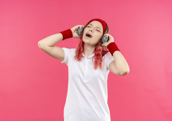 人戴着耳机 戴着头带的年轻运动女性 欣赏着她最喜爱的音乐 站在粉色的墙上 快乐地唱着一首歌享受歌曲最爱