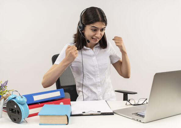 拳头穿着白衬衫 戴着耳机 手持麦克风 握紧拳头的年轻女子坐在桌子旁 把文件夹和笔记本电脑放在白墙上握紧姿势女人