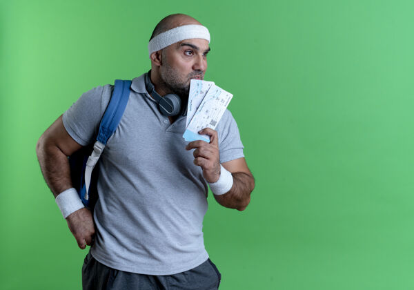 头带戴着头巾的成熟的运动型男人拿着两张机票 站在绿色的墙上困惑地看着一边教练运动员票