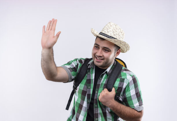 姿势穿着格子衬衫 戴着夏装帽子 背着背包的年轻旅行家站在白色的墙上愉快地挥手致意人人帽子
