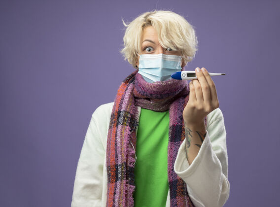 围巾生病的不健康的女人 短头发 戴着暖和的围巾 戴着防毒面具 惊慌失措地站在紫色的墙上 体温表在里面显示短裤不健康