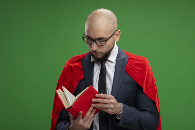 阅读超级英雄 留着胡子的商人 披着红斗篷 手里拿着打开的书 站在绿色的墙上看书立场举行男人